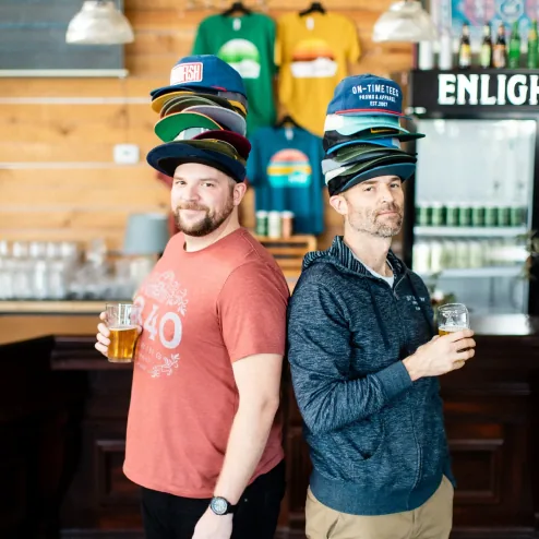 Joe and Ben of Craft Industry Apparel wearing stacks of custom hats in a taproom